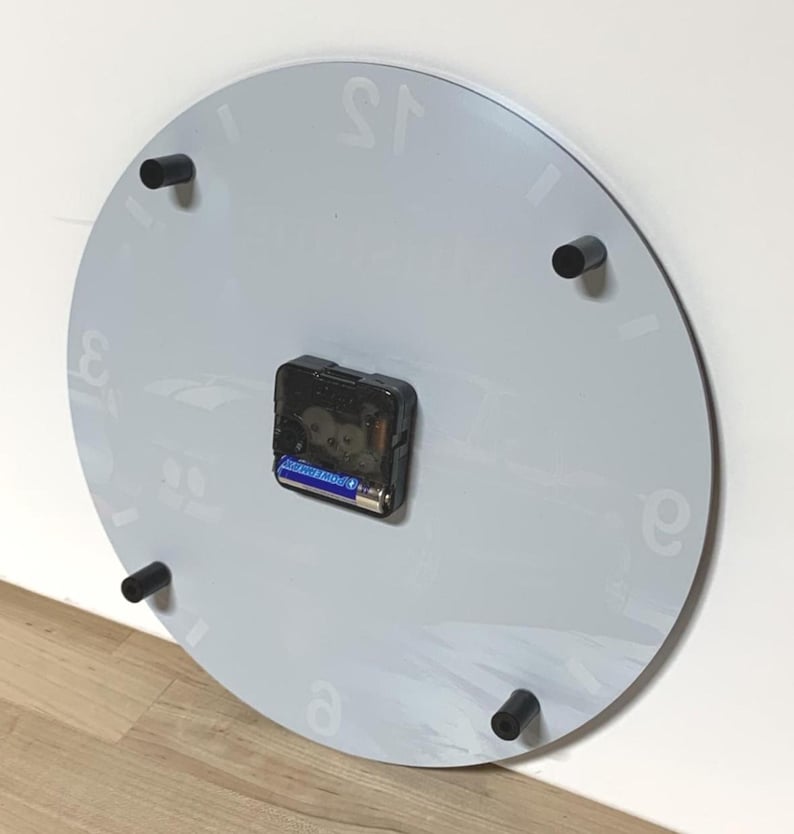 May include: A circular wall clock viewed from the back, featuring a light gray surface with faint numeral markings. A black plastic clock mechanism is centered on the back, holding a single AA battery. Four black cylindrical spacers are attached.