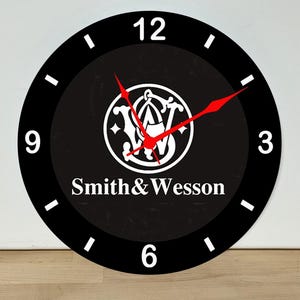 May include: A round wall clock with a black face and white numerals at the 3, 6, 9, and 12 o'clock positions. The center features the white Smith & Wesson logo and text. The clock has two thin, pointed red hands for indicating the time.
