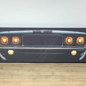 May include: A rectangular canvas wall art print featuring the front view of a dark-colored vintage muscle car. The design highlights a chrome bumper, a black grille, and four illuminated circular headlights glowing with a warm yellow light.