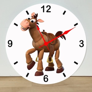 May include: A round wall clock with a white face, black numerals at 12, 3, 6, and 9, and black tick marks for other hours. The center features a brown cartoon horse with a saddle, and the clock has thin, bright red hour and minute hands.