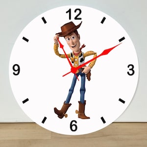 May include: A round wall clock with a white face, black hour markers, and bold black numerals at 12, 3, 6, and 9. The center features a colorful illustration of a cowboy character wearing a brown hat, yellow shirt, and blue jeans. Three bright red clock hands are positioned over the character.