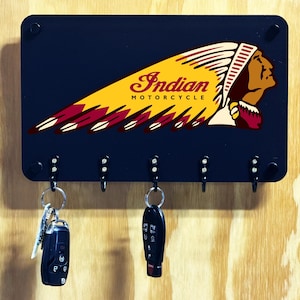 May include: A rectangular black wall-mounted key holder featuring the Indian Motorcycle logo with a yellow and maroon headdress graphic. The unit has five black hooks, with two sets of car keys hanging from the first and third hooks respectively.