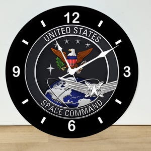 May include: A round wall clock with a black face and white numerals at 3, 6, 9, and 12. The center features the United States Space Command emblem, displaying a bald eagle, a globe, and a silver delta insignia with the text United States Space Command.