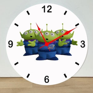 May include: A round white wall clock featuring three green three-eyed alien figures in blue space suits. The clock face has black hour markers and numerals at 3, 6, 9, and 12 o'clock. It includes three bright red clock hands for hours, minutes, and seconds.