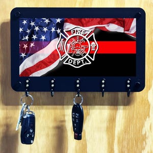 May include: A rectangular black wall-mounted key holder featuring a graphic of the American flag and a thin red line, centered with a white Fire Dept emblem. The unit has five black hooks, with two car key fobs hanging from the left and center hooks.