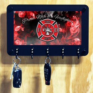 May include: A black rectangular wall-mounted key holder with five hooks. The center features a graphic with red smoke, floral patterns, a red and silver firefighter Maltese cross emblem, and the white script text Proud Wife of a Firefighter.