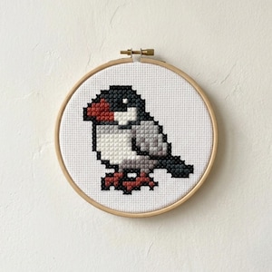 May include: A circular wooden embroidery hoop containing a cross-stitch pattern of a small bird. The bird features a black head, a red beak, a white chest, and gray wings. The design is stitched onto white Aida fabric with a visible grid texture.