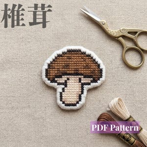 Puede incluir: Un parche de champiñón en punto de cruz con sombrero marrón y tallo crema, bordeado con hilo blanco. La imagen incluye tijeras de bordar doradas con forma de cigüeña y dos madejas de hilo, con el texto japonés 椎茸 y PDF Pattern.