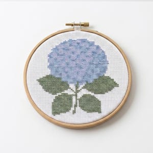 以下が含まれることがあります： 木製の円形刺繍枠に白い布が張られ、青いアジサイの花がクロスステッチで刺繍されています。花の部分は青と紫の様々な色合いで構成され、4枚の葉は落ち着いた緑色で刺繍されています。