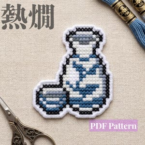 Peut inclure: Un écusson au point de croix représentant une bouteille de saké et une petite tasse bleu et blanc, avec les caractères japonais 熱燗 et le texte PDF Pattern. Le motif utilise des fils bleus, blancs et gris sur un fond en tissu blanc.
