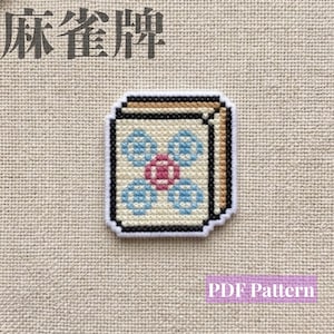 May include: A cross-stitch embroidery of a mahjong tile featuring a central pink flower surrounded by four blue floral elements on a cream background with black borders. The text "麻雀牌" is at the top and "PDF Pattern" is in a purple box at the bottom.