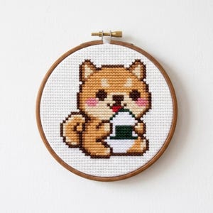以下が含まれることがあります： 木製の円形刺繍枠に、ピンクの頬をした薄茶色の柴犬が、白と緑の三角形のおにぎりを持っているクロスステッチ模様が施されています。デザインは白い布地の背景の中央に配置されています。