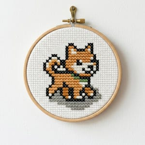 以下が含まれることがあります： 木製の円形刺繍枠に、緑色の首輪をしたピクセル調のオレンジと白の犬のクロスステッチ模様が施されています。犬は小さな灰色の地面の上に立っており、白い布地に黒い輪郭線で刺繍されています。