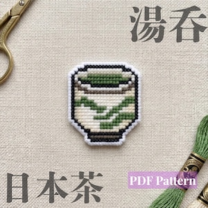 Puede incluir: Un diseño de punto de cruz de una taza de té japonesa con base blanca, patrones de hojas verdes y un borde marrón oscuro. El diseño está rodeado por un contorno negro. La imagen incluye tijeras doradas e hilo de bordar verde.