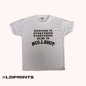 Könnte beinhalten: Ein weißes kurzärmeliges T-Shirt mit schwarzem Text auf der Brust, der DANCING IS EVERYTHING EVERYTHING ELSE IS BULLSHIT lautet. Das T-Shirt hat einen Rundhalsausschnitt und ein kleines Markenetikett im inneren Bereich des Kragens.