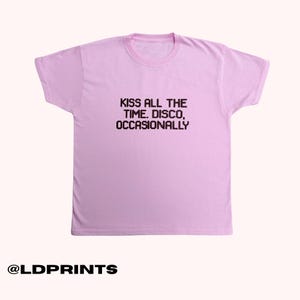 May include: A light pink short-sleeved t-shirt featuring black pixelated text centred on the chest that reads: KISS ALL THE TIME. DISCO, OCCASIONALLY. The garment has a crew neckline and a standard fit, presented against a plain light background.