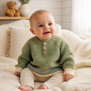 May include: A sage green long-sleeved knit sweater featuring a round neckline, three light-colored wooden buttons at the chest, and a ribbed hem with thin horizontal stripes in light blue and tan, paired with cream-colored ribbed knit leggings.