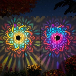 Peut inclure: Deux appliques solaires circulaires fixées sur une clôture, projetant des motifs de mandala multicolores vibrants dans des tons jaune, orange, bleu et violet sur la surface en bois, créant un affichage symétrique de lumière et d'ombre.