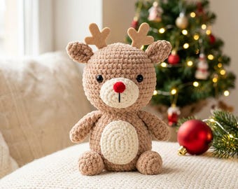 May include: A crocheted reindeer plush toy made of light brown yarn with a cream-colored belly and muzzle. It features small felt antlers, two black safety eyes, and a small red pom-pom nose. The toy has a round head and body.