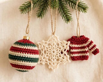 May include: Three hanging ornaments suspended from a pine branch: a spherical crochet ball with red, white, and green stripes, a white crocheted snowflake, and a miniature crochet sweater featuring horizontal red and white stripes.