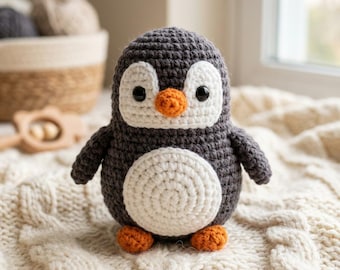 May include: A small, handmade crochet penguin toy featuring a dark gray body, a white face patch, a white belly, a small orange beak, and two orange feet. The penguin has two black plastic safety eyes and a textured, yarn-based construction.