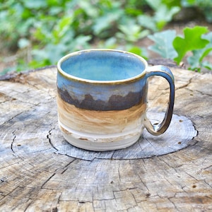 Può includere: Una tazza in ceramica con smalto a gradiente orizzontale, interno e bordo azzurro, una fascia centrale marrone scuro e una base color sabbia tendente al bianco sporco. La tazza presenta un manico curvo sul lato destro e una forma cilindrica.