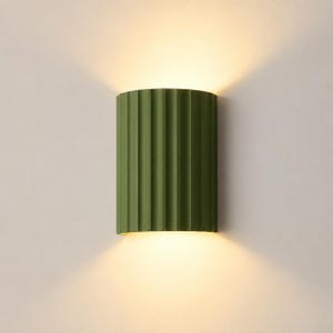 Pode incluir: Um candeeiro de parede semicilíndrico com uma textura canelada vertical num acabamento verde-azeitona mate. A luminária emite um brilho quente e suave pelas aberturas superior e inferior, projetando luz para cima e para baixo na parede.