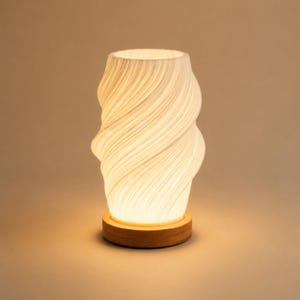 May include: A cylindrical table lamp with a white, translucent, spiraling textured shade that emits a warm glow. The lamp is supported by a circular, light-toned wooden base, creating a soft illumination that highlights the vertical ridges.