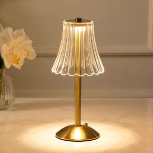 Può includere: Una lampada da tavolo con base in metallo dorato e stelo verticale che sostiene un paralume in vetro trasparente scanalato con bordo smerlato. La lampada emette una luce calda rivolta verso il basso, illuminando la superficie con un bagliore.