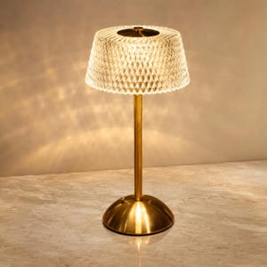 Op de afbeelding: Een tafellamp met een cilindervormige, gefacetteerde kap van helder acryl die een warme gloed uitstraalt. De lamp heeft een slanke, verticale metalen stang in messingkleur en een ronde, koepelvormige voet in messingkleur.
