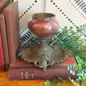 Könnte beinhalten: Ein Kerzenhalter im Vintage-Stil mit einer runden, irisierenden rotbraunen Glasschale auf einem verzierten, verwitterten messingfarbenen Metallsockel mit komplizierten Schnörkeln und blattartigen Details um den runden Fuß.