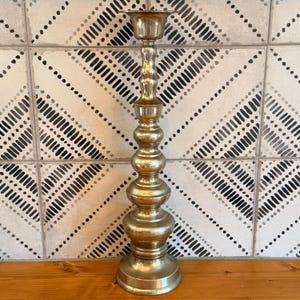 May include: A tall, polished brass candlestick holder featuring a series of stacked, rounded, and bulbous decorative sections along its central stem, topped with a wide, circular candle cup and supported by a sturdy, flared, circular base.