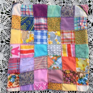 May include: A square patchwork quilt featuring a grid of multi-colored fabric squares. The textile squares display various patterns including floral prints, gingham checks, plaids, and solid colors, all joined together with visible whipstitch seams.