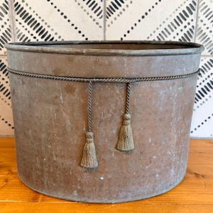 Puede incluir: Una maceta cilíndrica de cobre con una pátina desgastada y oxidada. El exterior presenta un borde de cuerda decorativo que rodea la parte superior, acentuado por dos borlas de cuerda colgantes en la parte frontal del recipiente.