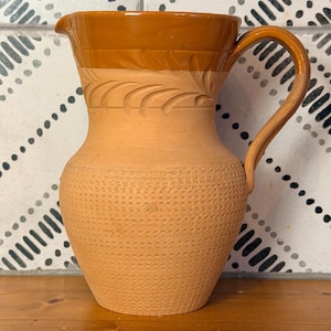 May include: A terracotta pitcher featuring a smooth, glossy amber-colored rim and handle. The upper neck displays a carved leaf pattern, while the rounded body is decorated with a textured, grid-like embossed design throughout.