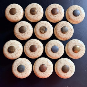 Puede incluir: Un conjunto de catorce pomos redondos para armarios con una base de cerámica de color crema con motas marrones y una pieza metálica circular central con acabados variados en bronce, cobre y latón oscuro.