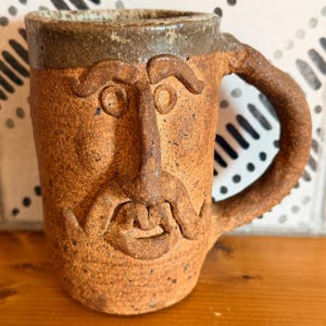 May include: A textured, brown ceramic mug featuring a sculpted face with prominent eyebrows, a long nose, and a mustache. The mug has a thick, curved handle and a darker, smooth rim at the top. The surface of the clay is speckled and rough.