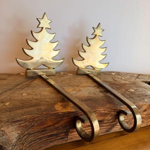 Könnte beinhalten: Zwei Strumpfhalter aus poliertem Messing, die jeweils eine stilisierte Weihnachtsbaum-Silhouette mit einem Stern an der Spitze zeigen. Die Metallhaken ragen nach vorne und enden in einer geschwungenen, kreisförmigen Form.