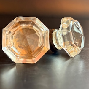 Peut inclure: Deux boutons de porte en verre transparent à facettes de forme octogonale. Chaque bouton présente un design géométrique à multiples faces qui capte la lumière, relié à une base métallique cylindrique sombre à l'arrière de la structure.