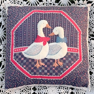 Puede incluir: Un cojín decorativo cuadrado con una aplicación central de dos gansos blancos sobre un fondo floral azul oscuro. El ganso de la izquierda lleva un pañuelo rojo y el de la derecha una pajarita azul. El diseño está enmarcado por un borde octogonal rojo.