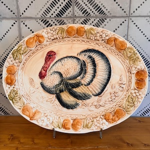 Puede incluir: Una bandeja de servicio ovalada de cerámica que presenta un pavo en relieve tridimensional en el centro, con una carúncula roja y plumas de puntas oscuras. El borde está decorado con un patrón en relieve de peras, uvas y hojas en tonos crema y beige.