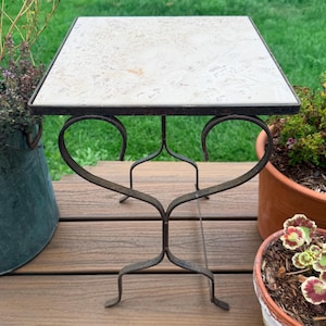 May include: A square side table featuring a light beige, speckled stone top surface set within a dark brown metal frame. The base is constructed from curved, heart-shaped metal supports that connect to four feet, providing a stable foundation.