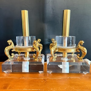 Puede incluir: Un par de lámparas de mesa con bases cuadradas de acrílico transparente, vástagos cilíndricos centrales de latón y detalles decorativos de metal dorado con forma de cabezas de animales estilizadas y patas curvas.