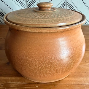May include: A round, terracotta-colored ceramic jar with a matching lid. The jar features a smooth, bulbous body and a textured, circular pattern on the top of the lid, which includes a small, centered knob handle for easy removal and secure closure.