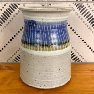 May include: A ceramic vase with a flared rim and a cylindrical body. The upper section features vertical blue stripes over a white base, separated by a dark brown band from the lower section, which is a speckled light gray with horizontal ridges.
