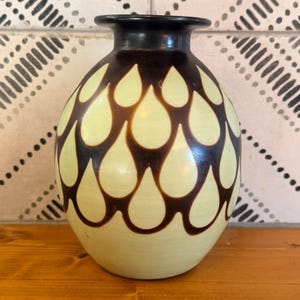 May include: A rounded ceramic vase with a smooth finish, featuring a cream-colored base and a repeating dark brown teardrop or petal pattern that covers the body. The vase has a short, narrow neck and a wide, flat, dark brown rim.