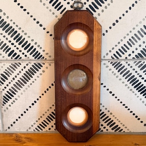 May include: A vertical, octagonal wooden wall plaque featuring three circular, recessed indentations. The top and bottom indentations contain smooth, light-colored circular inserts, while the center indentation holds a textured, brownish-gray circular insert. A small metal hanging loop is attached to the top.