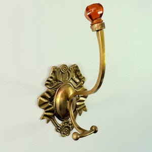 May include: A wall-mounted brass coat hook featuring an ornate, bow-shaped backplate with a central oval medallion. The hook has a curved arm extending upward, topped with a faceted, translucent amber-colored glass knob and a smaller lower hook.