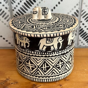 May include: A cylindrical ceramic jar with a removable lid featuring a sculpted elephant handle. The exterior is decorated with a black and cream geometric pattern and a repeating motif of elephants in profile around the center of the vessel.