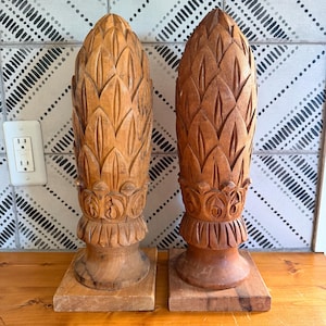 May include: Two tall, light brown wooden finials carved in a pointed, leaf-like pattern. Each finial features a textured, repeating scale design that tapers to a point, resting on a circular base attached to a square wooden block stand.